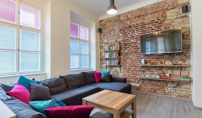 Common room with l-couch, exposed brick walls, TV, and bookshelves with books and games at The RoadHouse hostel in Prague.
