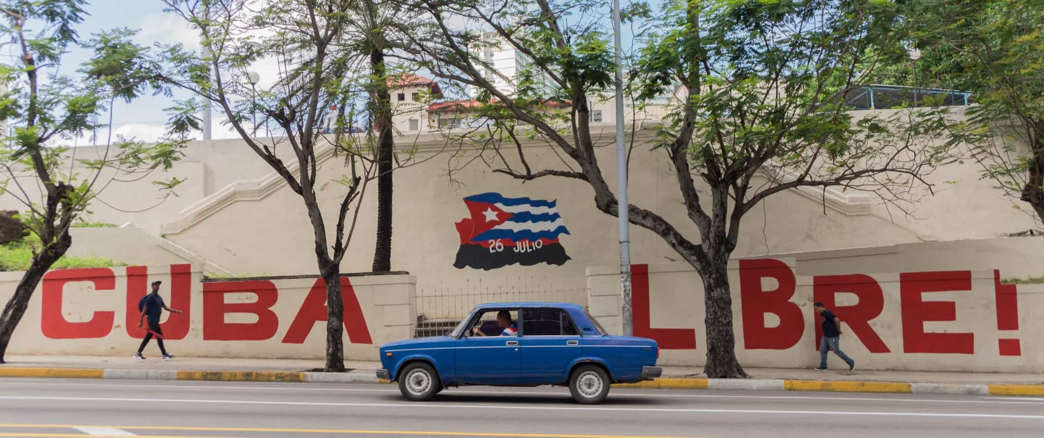 在古巴哈瓦那，一辆蓝色的旧汽车行驶在街道上，经过一幅写着“古巴自由”和古巴国旗的壁画