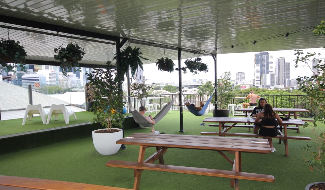 在澳大利亚布里斯班的城市背包客酒店，屋顶露台上有野餐桌，人们在吊床上闲逛。
