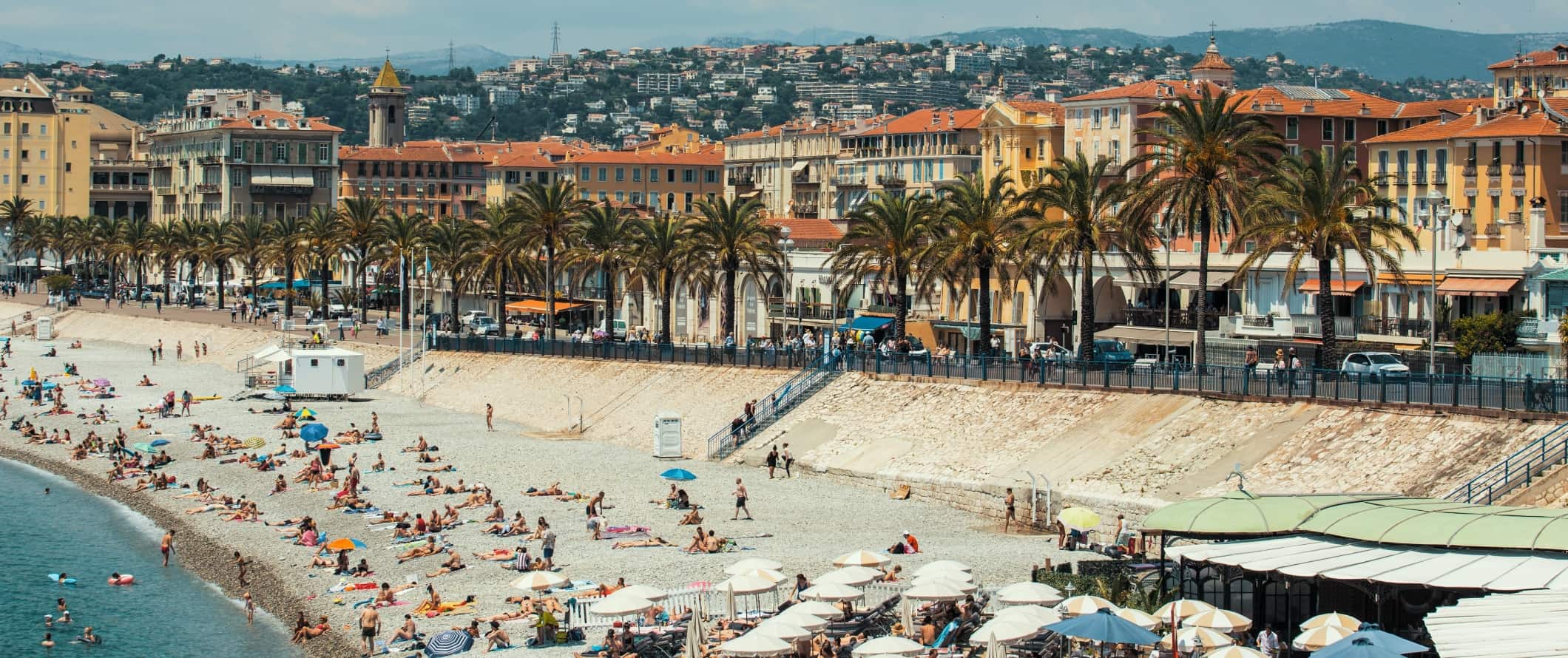 人们躺在沙滩上的棕榈树lined promenade with the city of Nice, France rising in the background