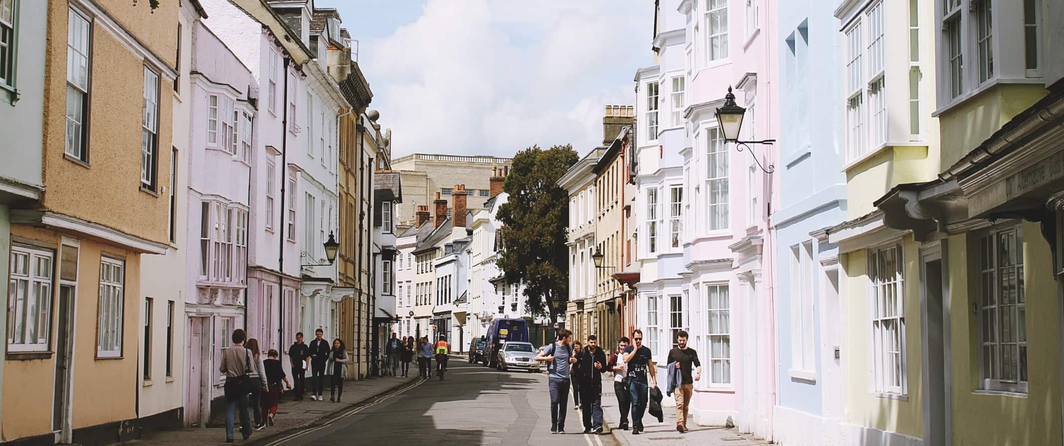 People walking down a street lined with pastel-colored townhouses in the town of Oxford, England