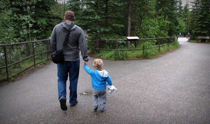 A dad and toddler walking through a park hand in hand while on vacation