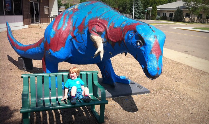 A baby crying while sitting on a bench near a blue a dinosaur statue while on vacation