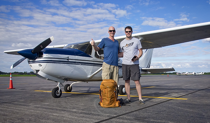 Two guys posing in front of an airplane with a backpack and a thumbs up
