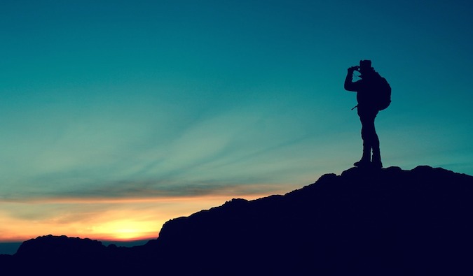 the silhouette of a backpacker standing on a cliff at sunset