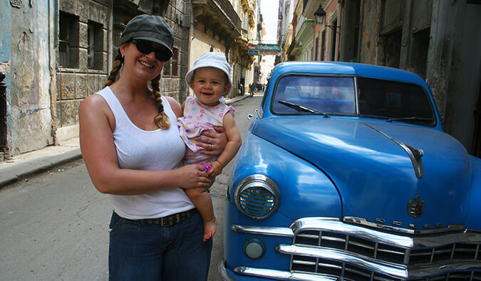 古巴一名妇女抱着一个婴儿，旁边是一辆蓝色的老古董车