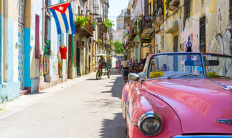 一辆经典的粉色汽车行驶在哈瓦那五颜六色的街道上，背景是古巴国旗