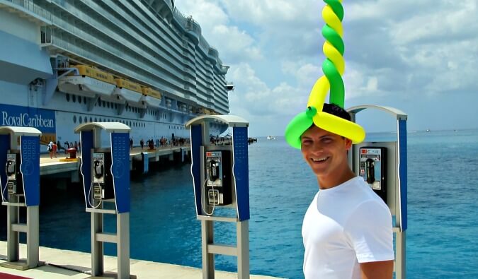 游牧的马特戴着一顶气球帽，在一个港口拍照，左边是皇家加勒比游轮的背景