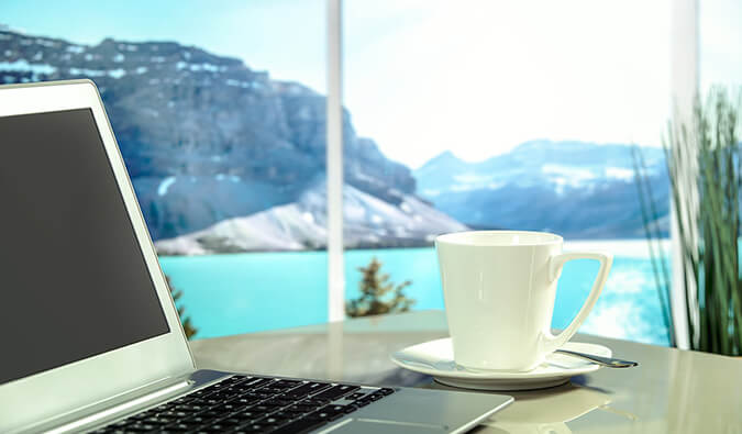 桌上放着笔记本电脑、咖啡杯和茶托，旁边的窗户可以看到远处的湖泊和群山