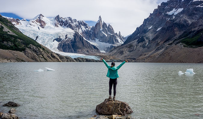 一名身穿绿色夹克的女子站在被雪山环绕的湖中的一块岩石上