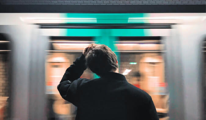 man scratching his head standing on a platform with a train passing by