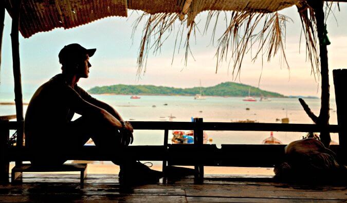 一个人的轮廓部分坐在一个小木屋里，他的背包在前景。背景是沙滩船和大海